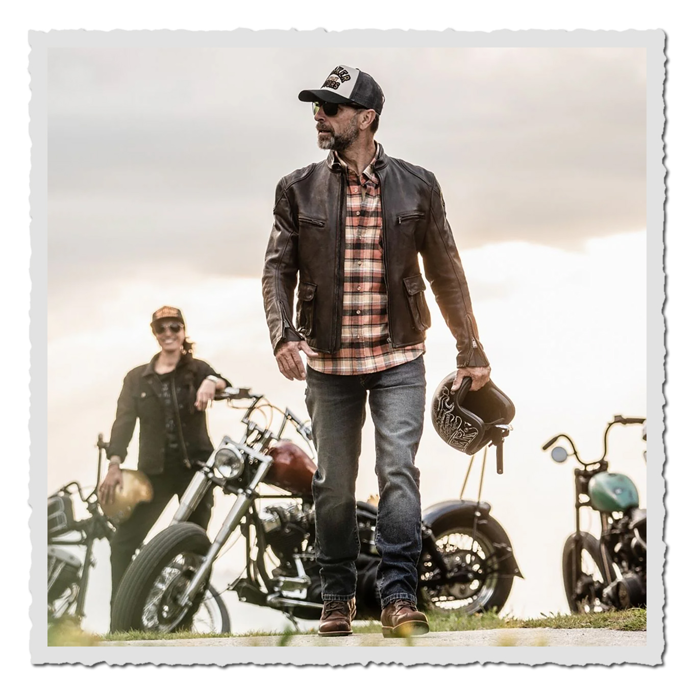 Mann trägt dunkelblaue Jeans, braune Lederjacke und Kappe vor mehreren Motorrädern im Freien.
